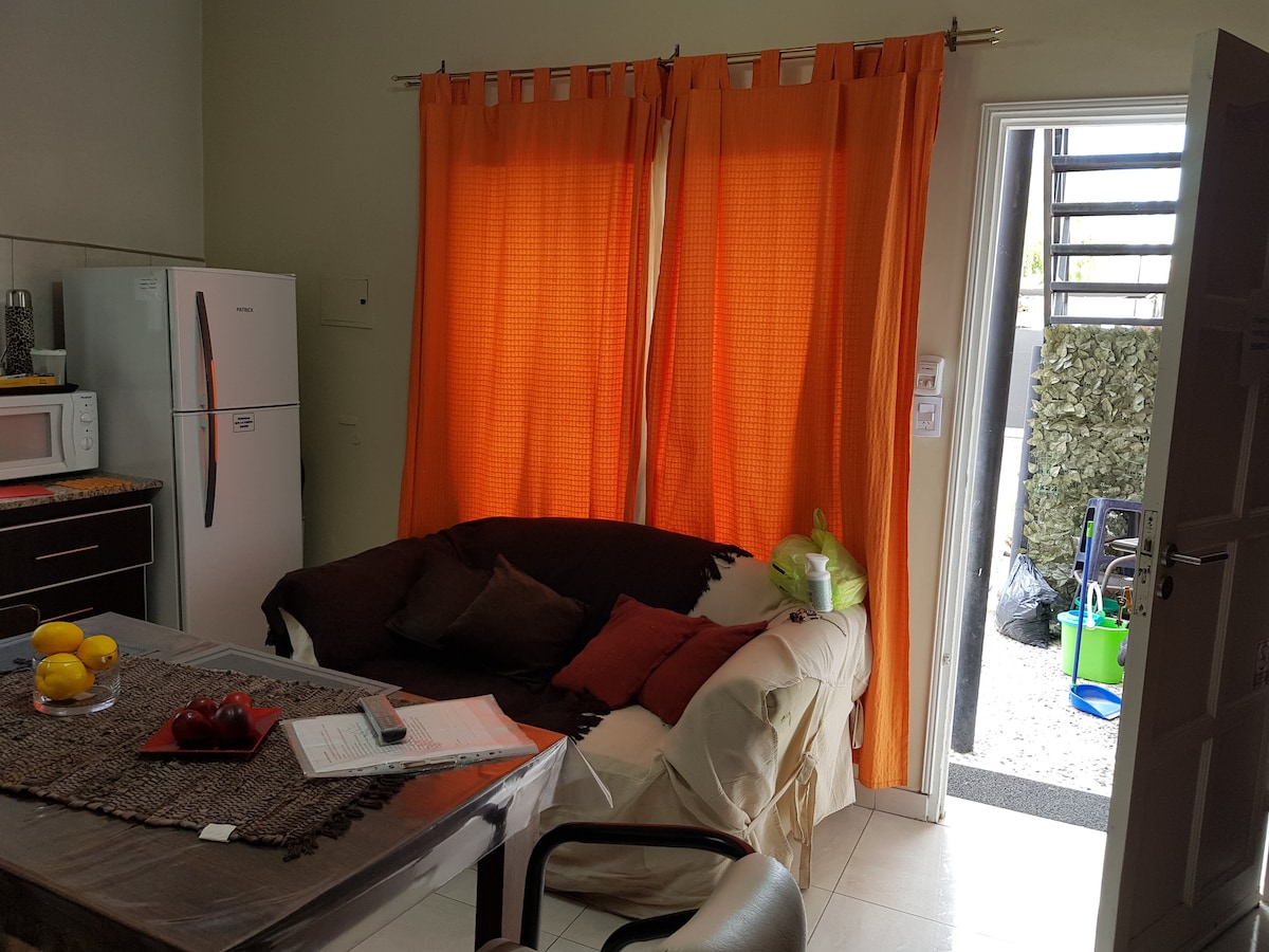 A cozy living space features an orange curtain draping over a doorway. A light-colored sectional sofa is complemented by dark cushions. A wooden table with a decorative runner sits nearby, and a kitchen area with a refrigerator and microwave is visible, enhancing the functionality of the room.