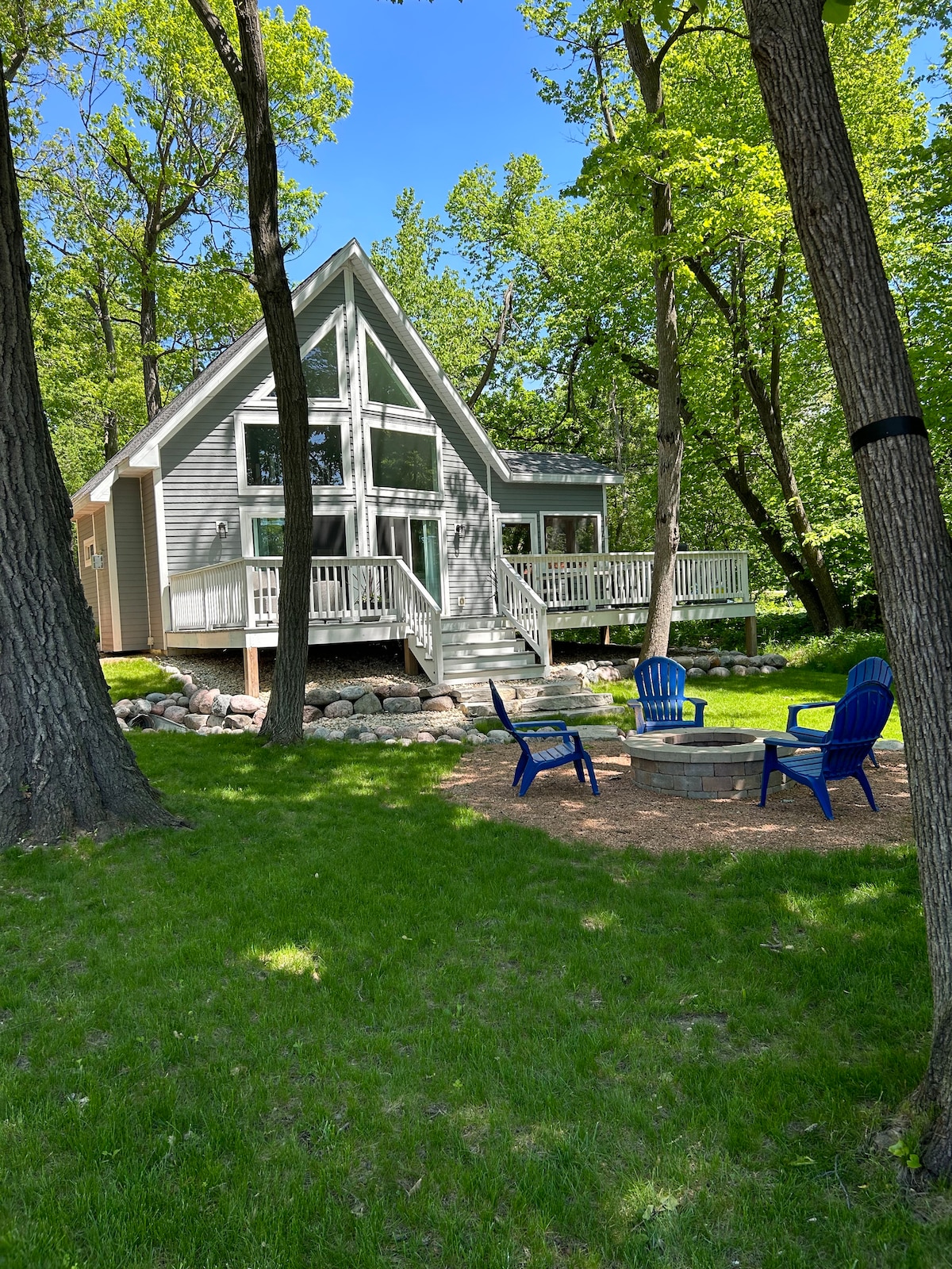 A modern A-frame house is surrounded by tall trees in a natural setting. The front deck is visible, leading to a spacious outdoor area with blue chairs arranged around a fire pit. Lush green grass blankets the ground, providing a serene atmosphere.