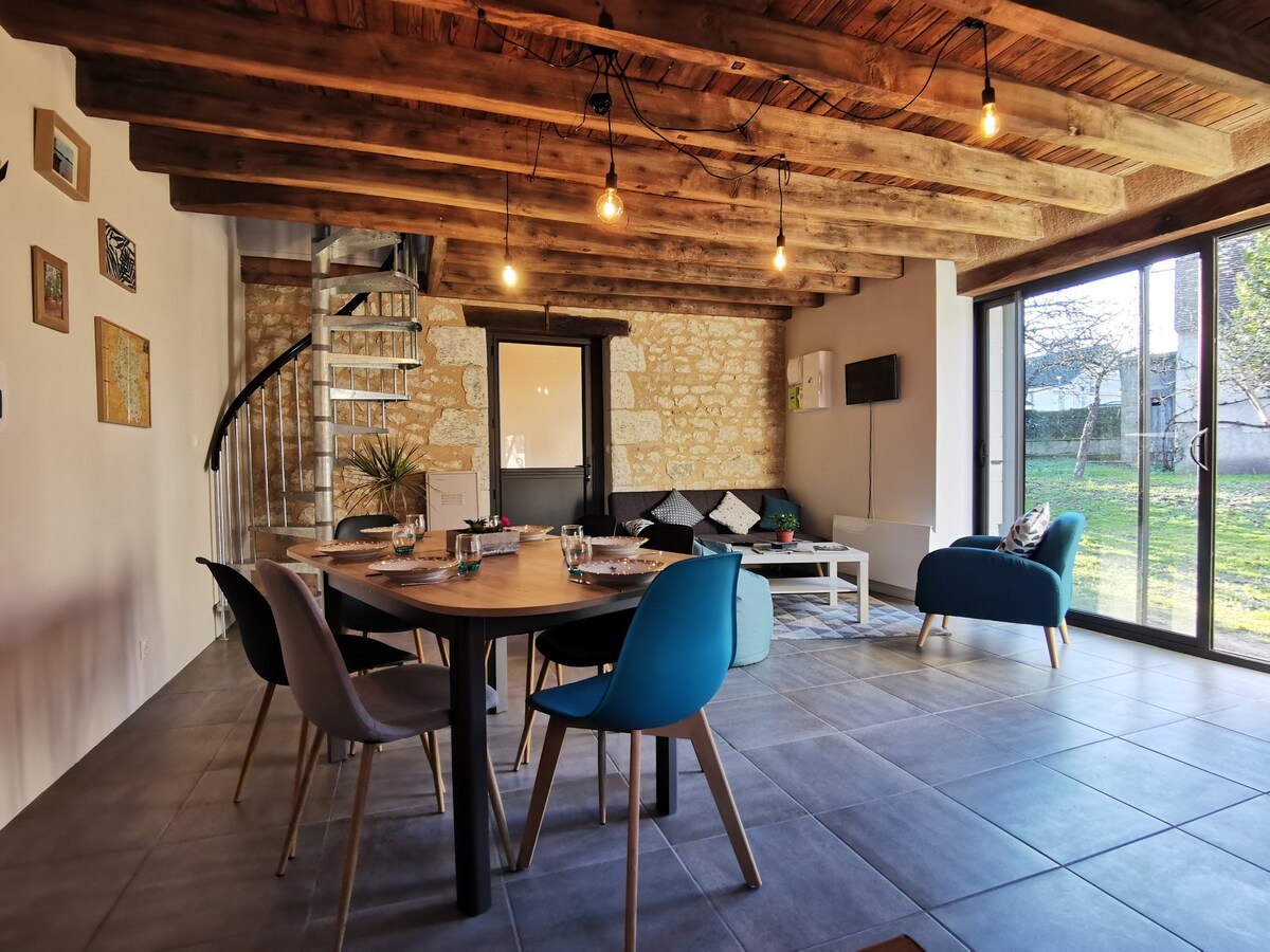 The living area features a circular dining table surrounded by modern chairs. A cozy seating arrangement includes a dark sofa and a blue accent chair. Natural light filters through large glass doors, illuminating the space and providing a view of the outdoor greenery.
