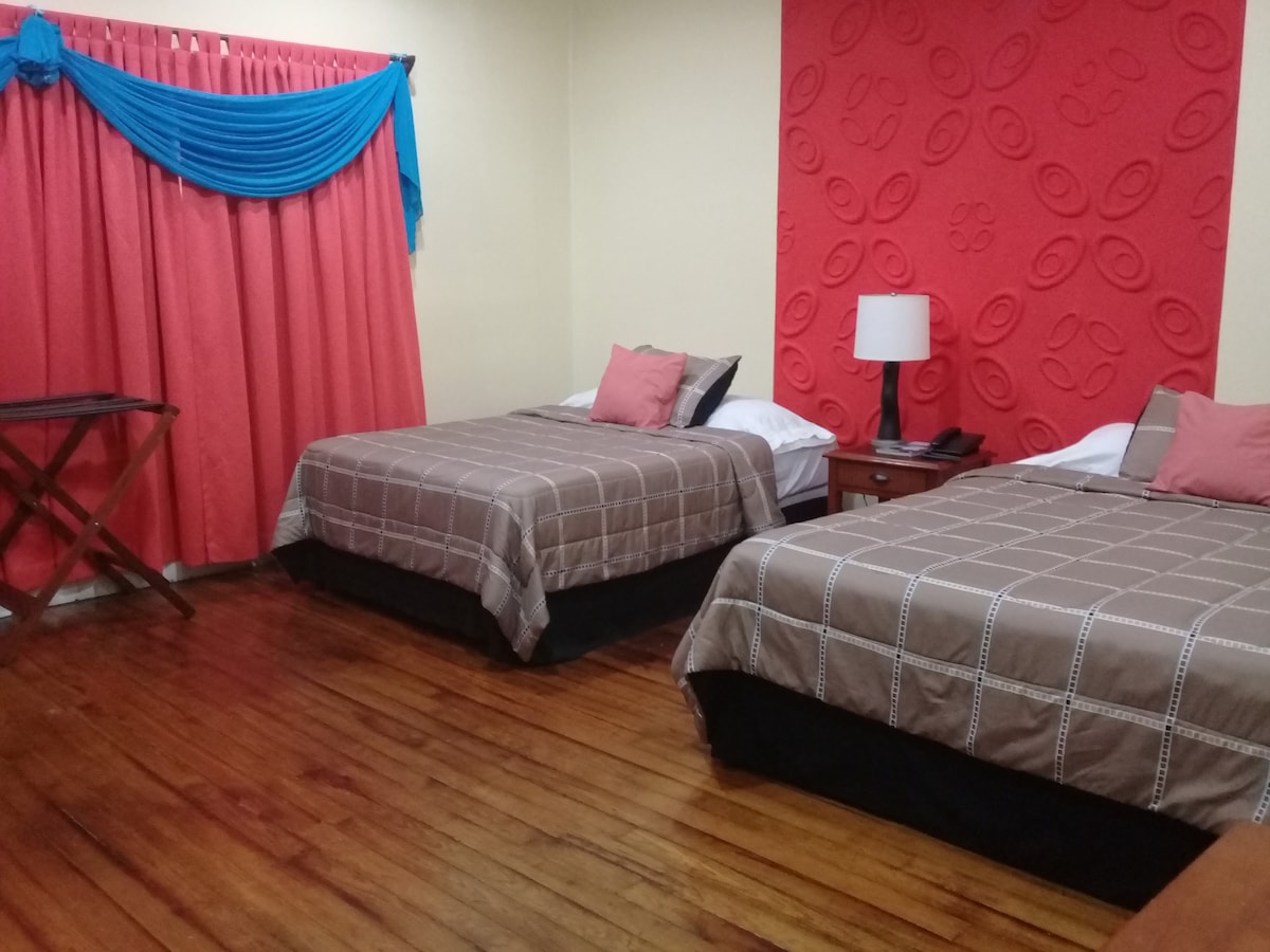 The room features two spacious beds with neutral bedding, complemented by orange and pink decorative pillows. A vibrant red wall adds color, while a blue curtain drapes over a window. A bedside table with a lamp is positioned next to each bed, and a folding table is visible.
