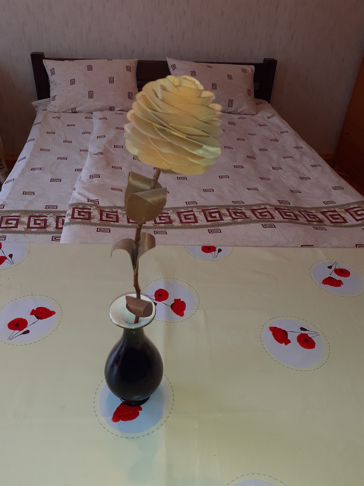 A simple room is presented with a spacious bed adorned with patterned bedding. A table is set with a pale yellow tablecloth featuring poppy designs. A single flower in a dark vase serves as a focal point in the center of the table.