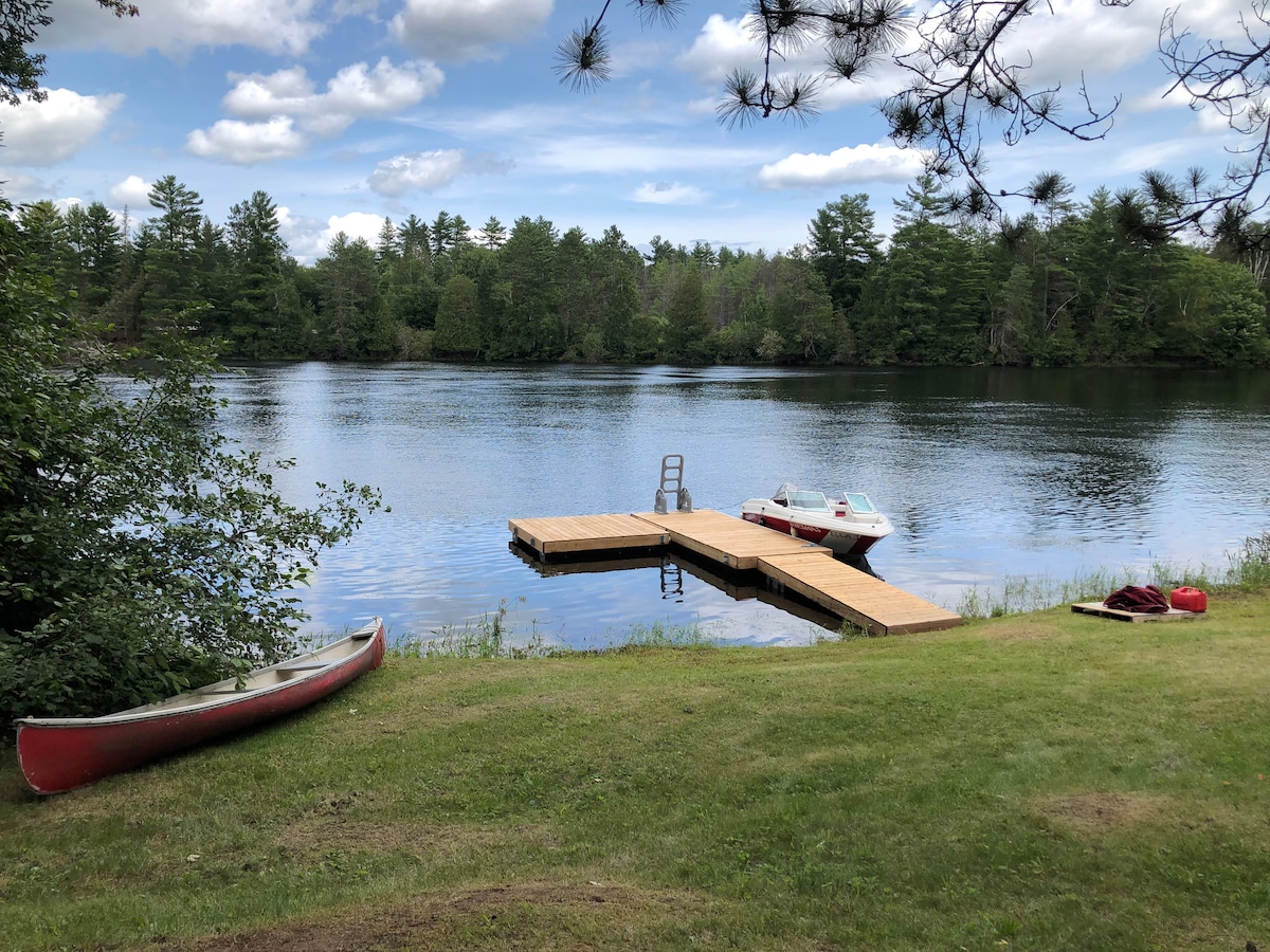 A wooden dock reaches out into the calm waters of the Madawaska River, accompanied by a canoe and a small boat nearby. Lush greenery surrounds the water, with trees lining the riverbank under a bright blue sky dotted with clouds.