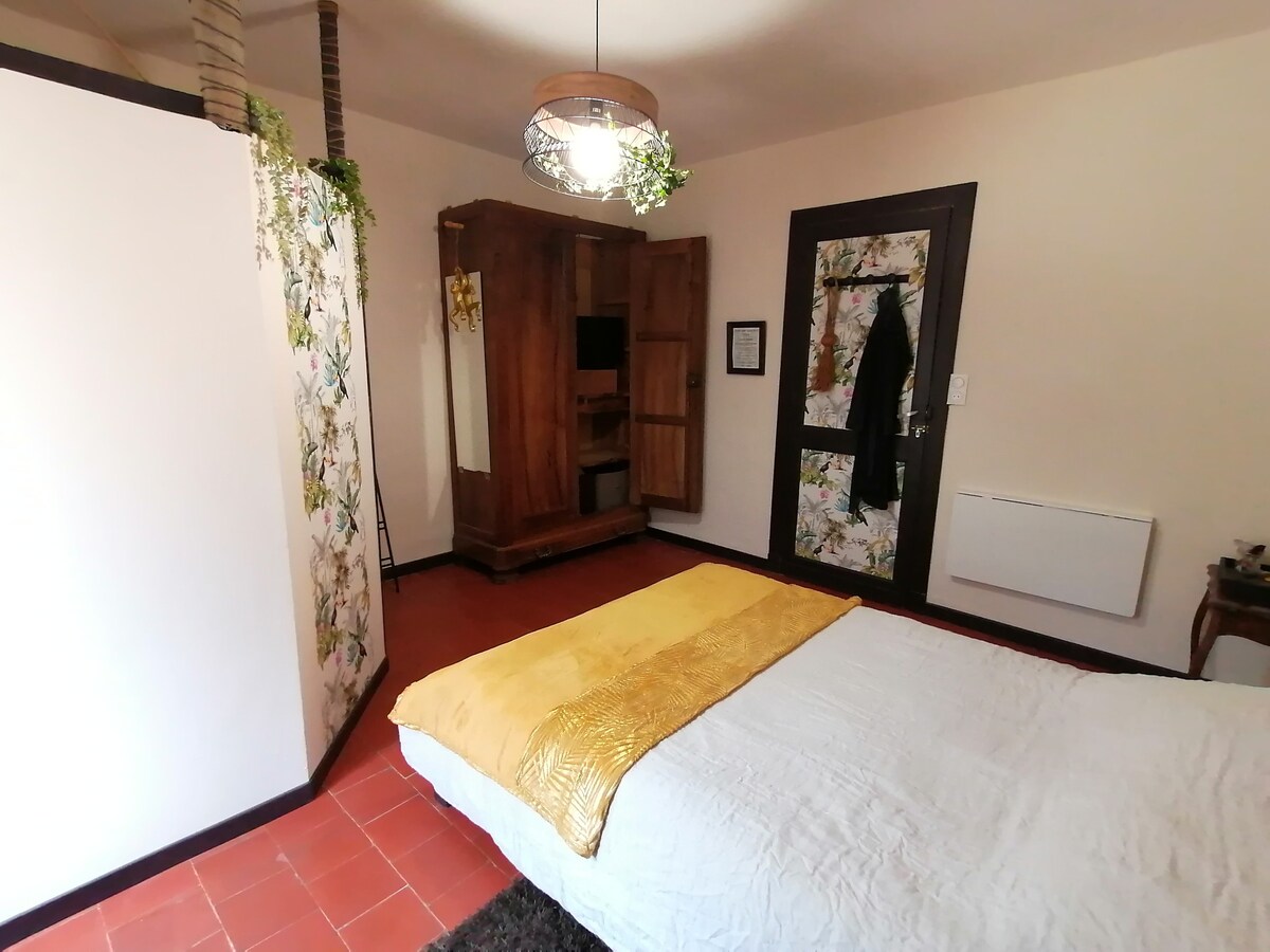 A cozy bedroom is presented with a spacious bed draped in white linens and a golden throw. A wooden wardrobe stands against the wall, while a decorative partition with floral accents adds privacy. Light fixtures illuminate the inviting space, creating a sense of warmth.
