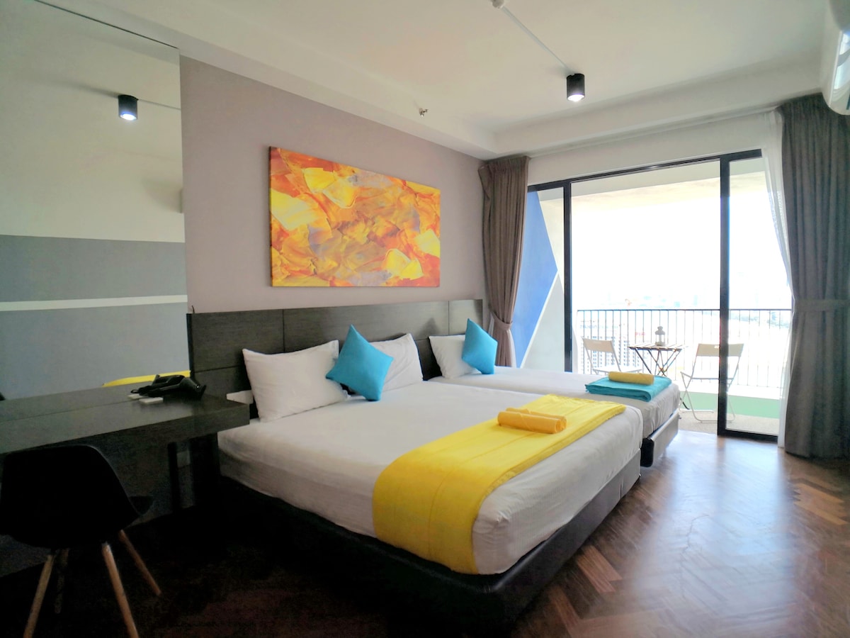 A spacious studio room is displayed with two beds, finished in calming tones. Bright blue pillows and a towel add color to the bedding. A modern work desk is seen beside a glass door, leading to a private balcony with views of the surroundings.