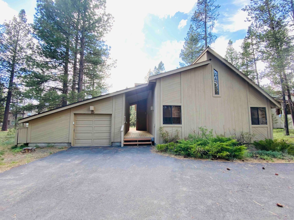 The exterior of a modern home is displayed among tall pine trees. A sloped roof and large windows characterize the structure, while a paved driveway leads to a garage. A wooden pathway leads to the entrance, framed by green landscaping.