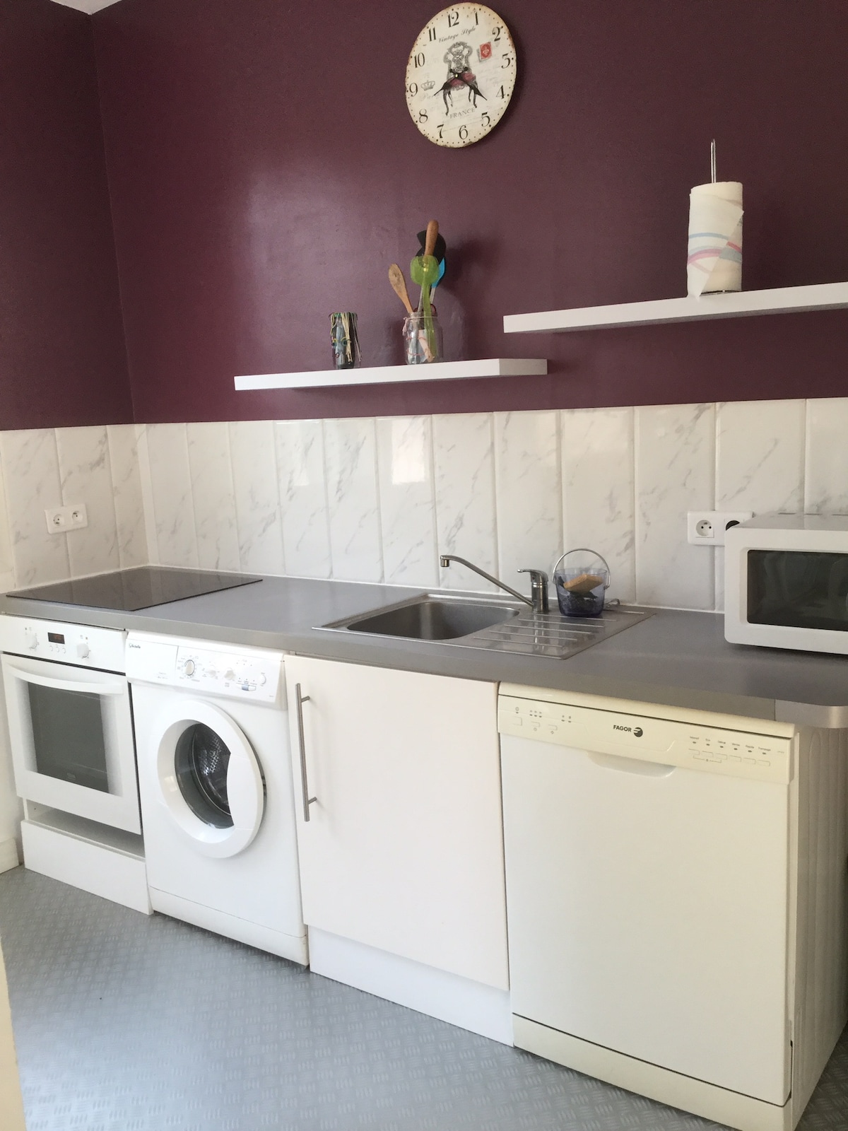 A functional kitchen area is equipped with a washing machine, dishwasher, and a combination oven with a microwave. A stainless steel sink is situated between the appliances, with two shelves above displaying kitchen utensils. A clock on the wall adds a practical touch.