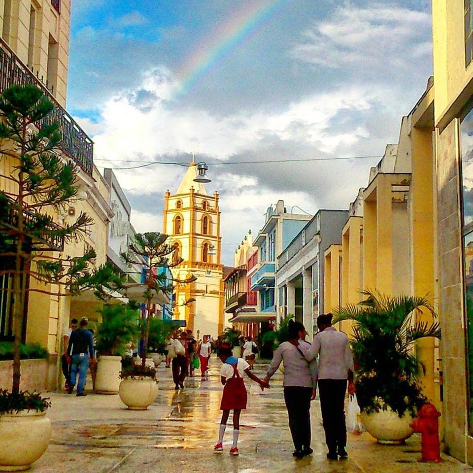 A vibrant street scene in downtown Camaguey features people walking along a bustling promenade lined with colorful buildings. The iconic church tower is seen in the background, framed by a rainbow. Potted plants add greenery to the lively atmosphere.