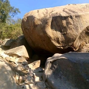 Large natural rocks are prominently featured, showcasing a rugged texture and earthy tones. The surrounding area contains smaller boulders, with shadows cast by the trees above, creating a serene and raw landscape. A pathway is visible, leading through the rocky terrain.