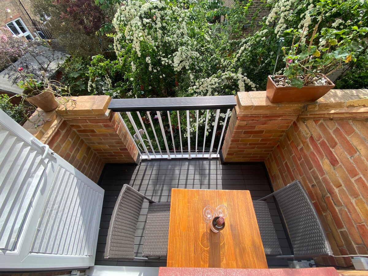 A balcony area is showcased, featuring a small wooden table and two woven chairs. Potted plants sit on the railing, overlooking lush greenery that fills the view. Light filters through the surrounding foliage, providing a serene outdoor space.