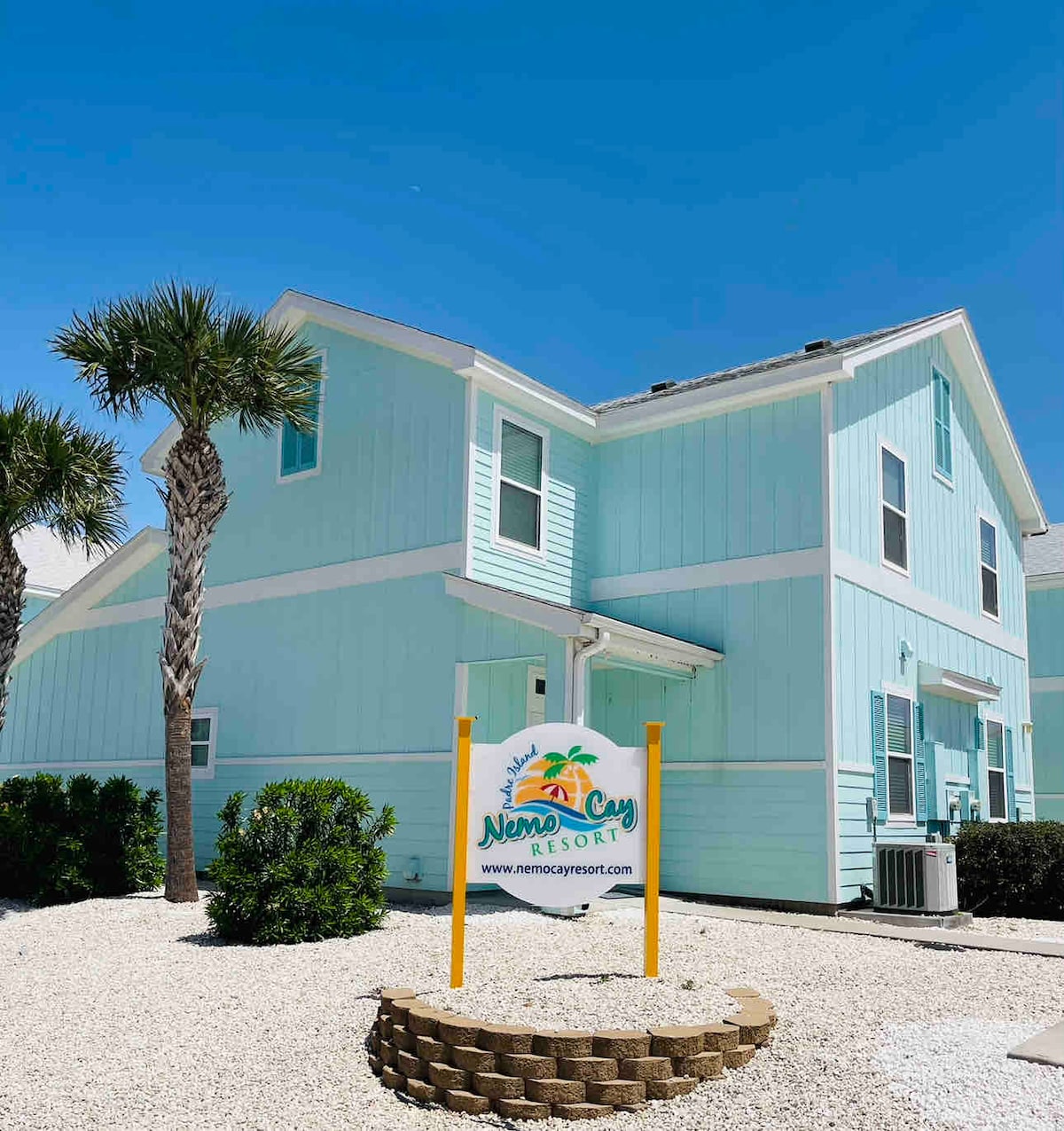 The exterior of the Nemo Cay Resort features a light blue building with a sign prominently displayed in front. Lush palm trees and well-maintained greenery surround the structure, while a clear blue sky highlights the inviting atmosphere of the resort.