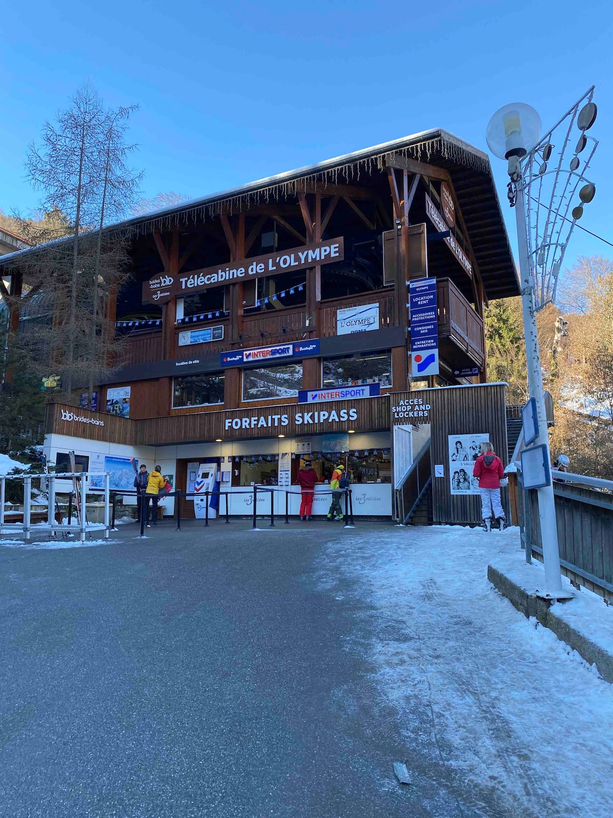 The exterior of the Télécabine de l'Olympe ski lift is shown, featuring a wooden structure with multiple windows and signage indicating ski passes. Snow covers the ground, and several people are present, dressed for cold weather.