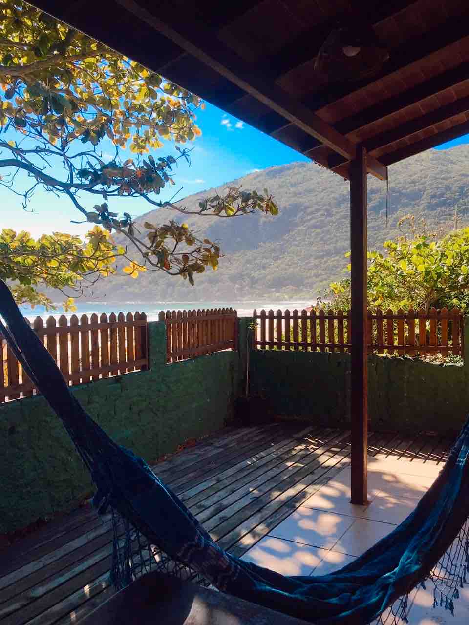 A shaded outdoor space is presented, featuring a hammock suspended beneath a wooden roof. The area is framed by a rustic fence, while lush greenery and a distant mountain can be seen in the background, suggesting a peaceful environment.