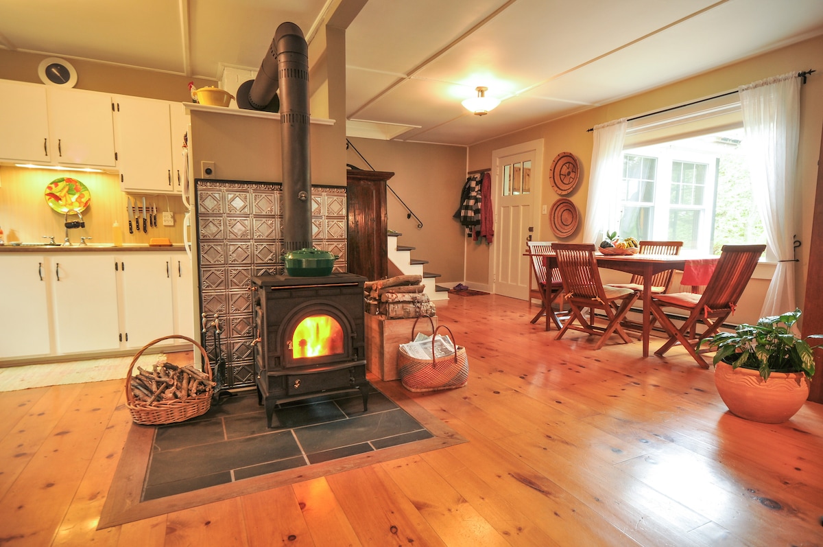 A warm and inviting living space features a stylish wood stove providing warmth in the center. The rustic wooden floor complements the light-colored walls. To the side, a well-equipped kitchen is visible with white cabinetry. A dining area with wooden chairs is illuminated by natural light from a nearby window.