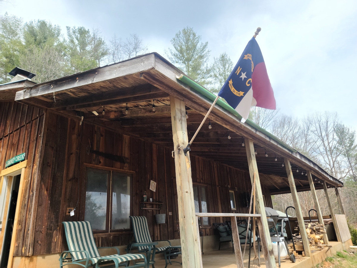 The cabin's porch features two striped lounge chairs, a North Carolina flag, and a rustic wooden structure. Natural wood tones and large windows provide a welcoming facade. A comfortable outdoor space is created under a covered area that enhances relaxation in the surrounding nature.