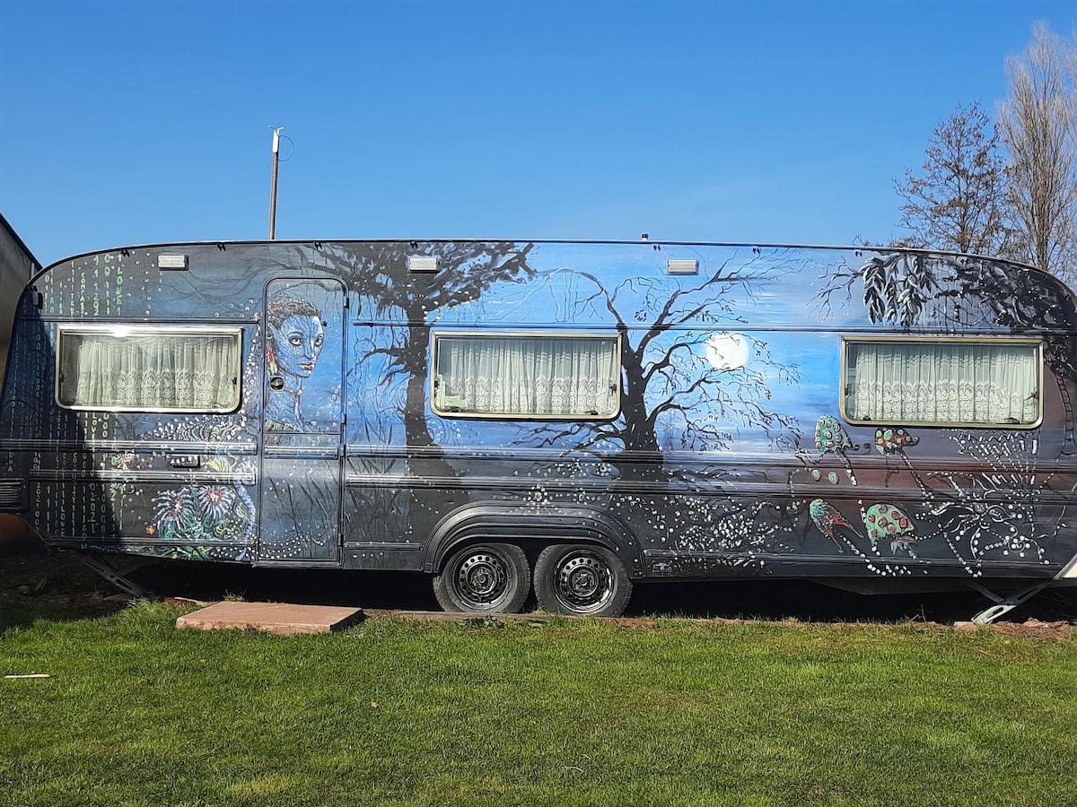 An older caravan with a striking mural adorns its exterior, depicting serene landscapes and whimsical figures. Bright blue skies are visible above, while lush green grass surrounds the base of the caravan. Large windows offer glimpses of natural light shining through.