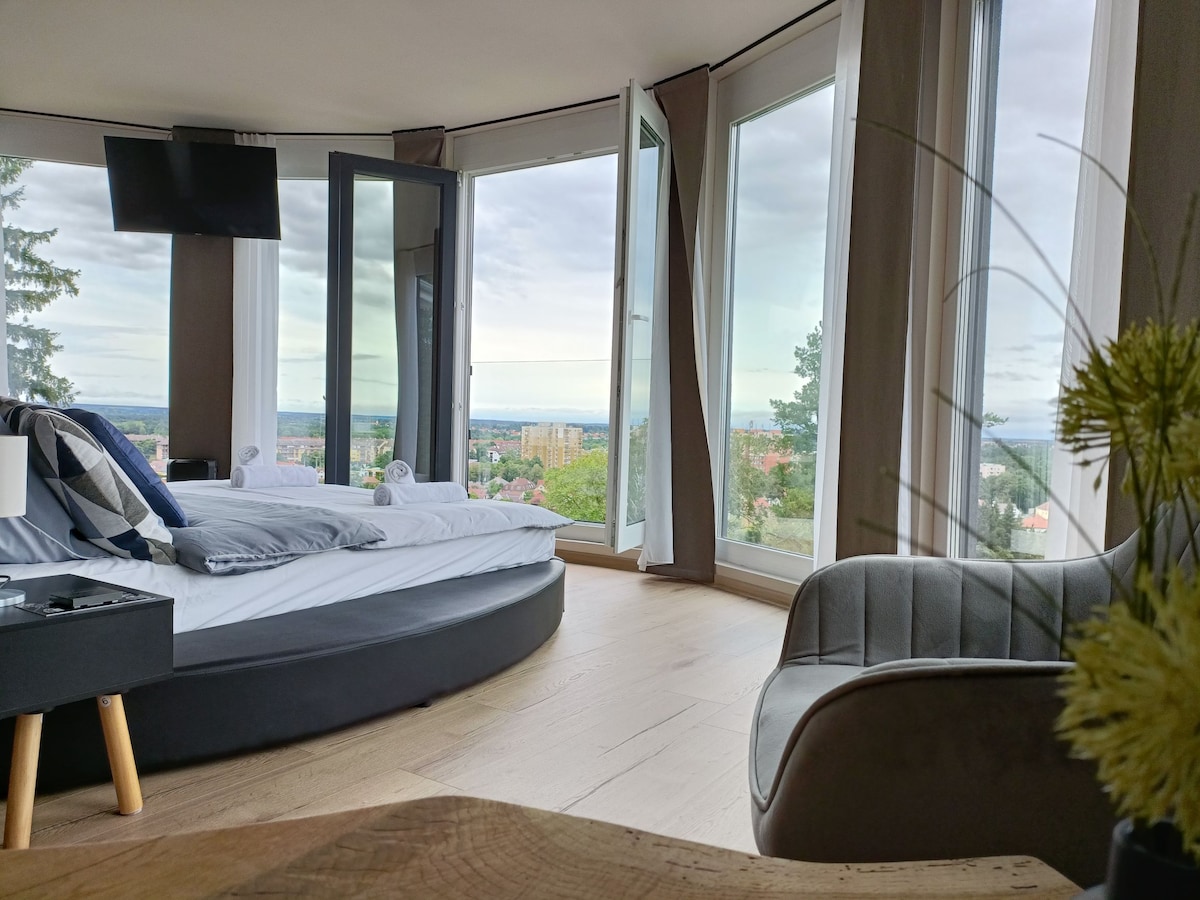 A spacious room features large windows that offer panoramic views of the surrounding landscape. A rounded bed is centered in the room, complemented by a minimalist television on the wall. A cozy chair is positioned near a unique wooden table, enhancing the inviting layout.