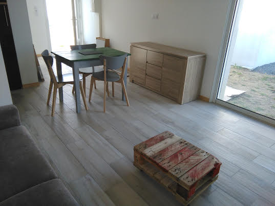 A modern studio space features a dining area with a rectangular table and four chairs. A light wood sideboard provides storage, while a casual seating area includes a sofa. Large glass doors allow natural light and open to a small outdoor space.