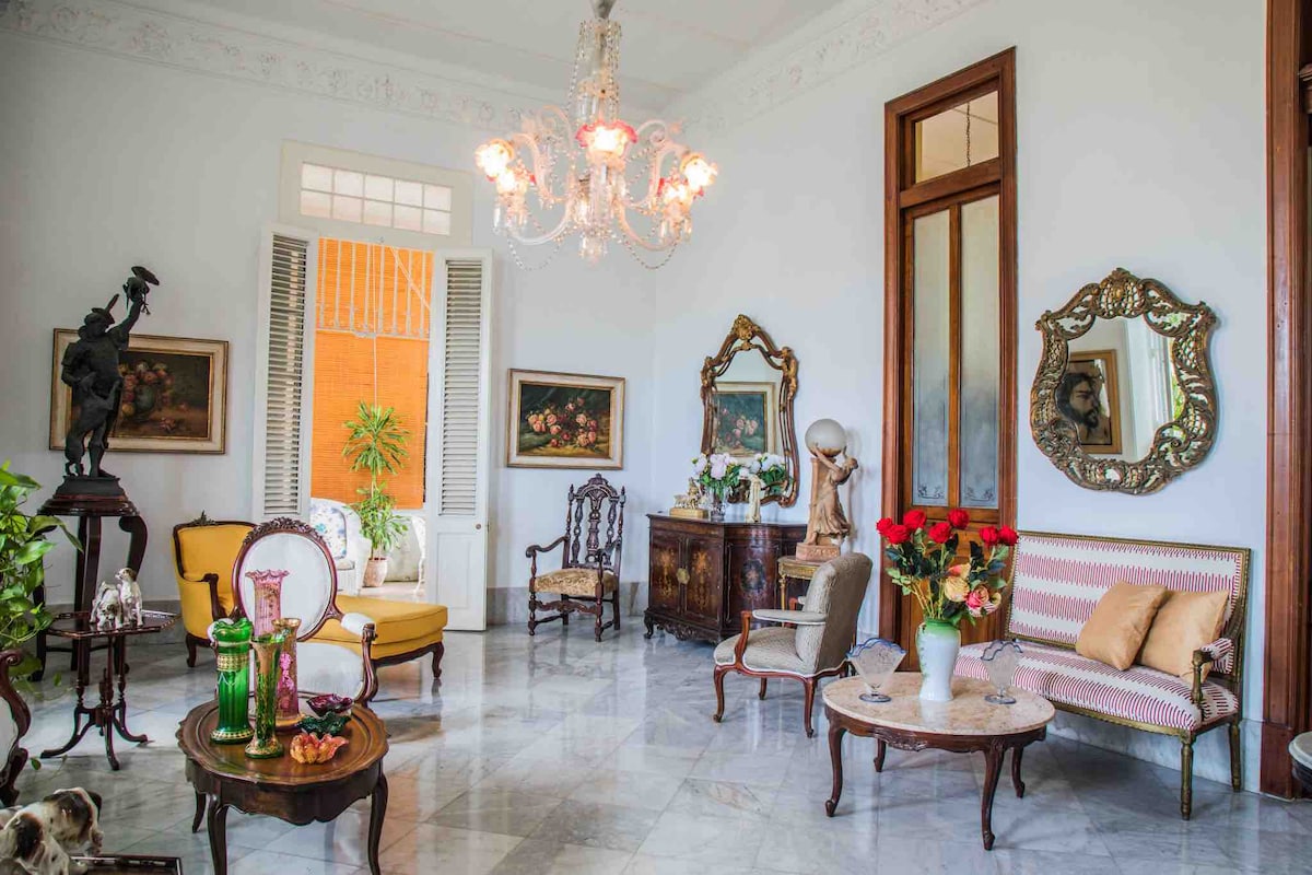 An elegant living room features a chandelier overhead, complementing ornate mirrors and decorative artwork. Various seating arrangements include a vintage sofa and armchairs, while a wooden table displays fruit and decorative items. Large windows allow natural light to enhance the bright atmosphere.