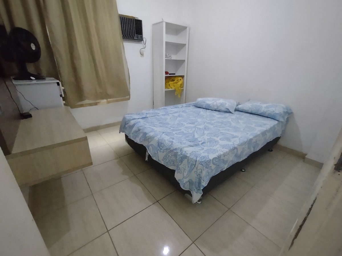 A simple bedroom is shown featuring a spacious bed with light blue bedding. A small fan is positioned nearby, and natural light enters through a window with beige curtains. A shelf is present against the wall, providing additional storage.