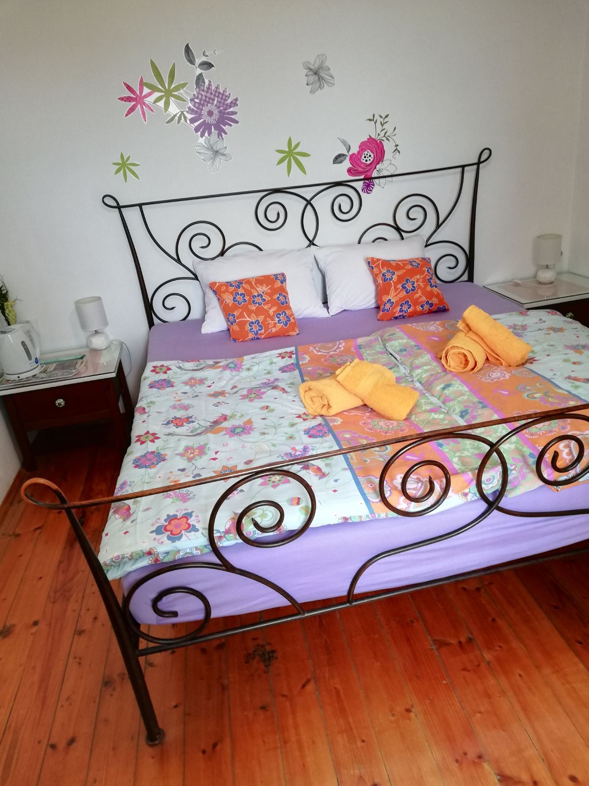 A cozy bedroom showcases an ornate metal bed with a colorful bedding set, complemented by bright decorative pillows. The walls feature floral accents, and bedside tables hold lamps. A calming atmosphere is created by the light wooden flooring and soft tones in the decor.