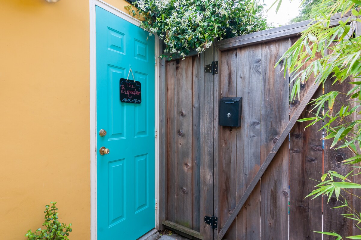 A vibrant turquoise door invites guests into the bungalow, framed by green foliage and a wooden fence. A simple sign hangs on the door, while a nearby mailbox completes the charming entrance.