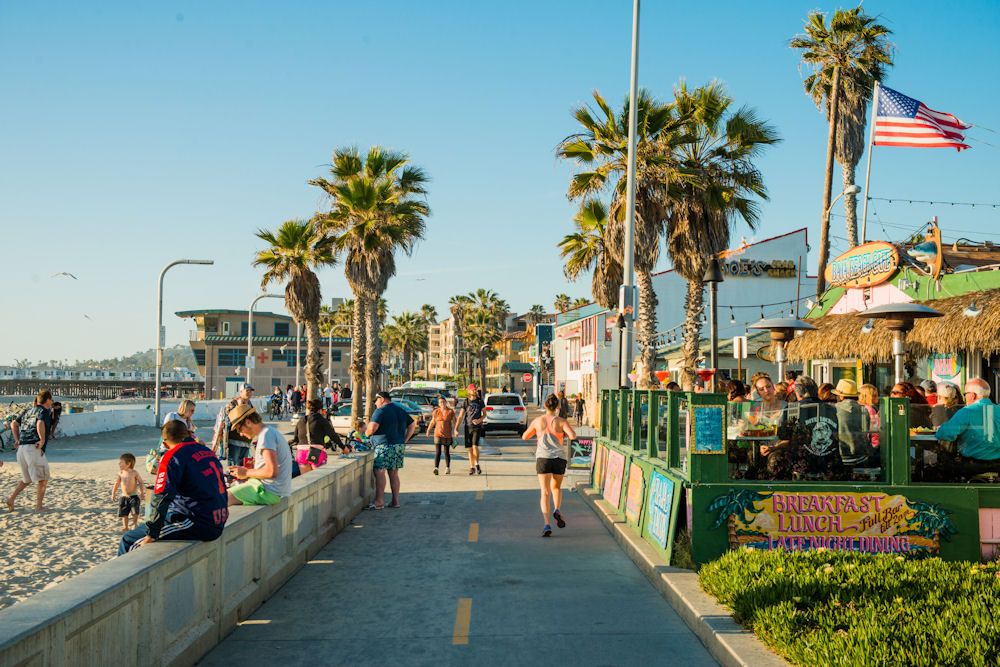 A lively beachfront scene features a wide pathway lined with palm trees and people. To the left, beachgoers relax on the sand, while a colorful eatery is visible nearby, showcasing vibrant signs. An American flag waves overhead, adding a patriotic touch to the coastal atmosphere.