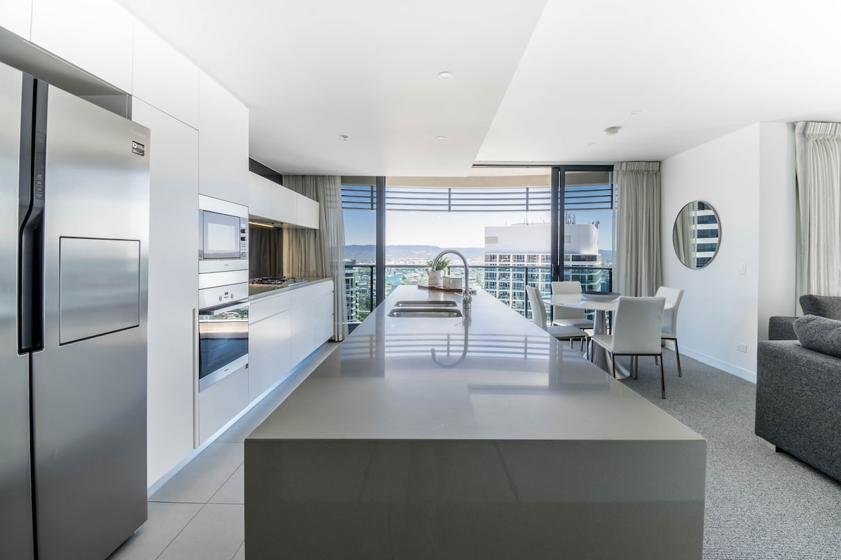 A modern kitchen is displayed with sleek cabinetry and stainless-steel appliances. A spacious grey island countertop provides ample preparation space, while large windows offer views of the city skyline. A round dining table with chairs is positioned nearby.