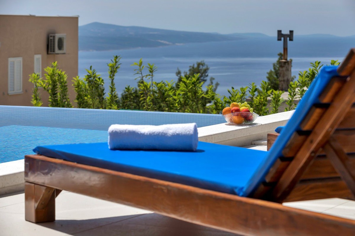 A sun lounger with a blue cushion is positioned beside a serene pool, offering a view of the sea and mountains in the background. A bowl of fruit sits on a nearby table, accompanied by a rolled towel resting on the lounger.