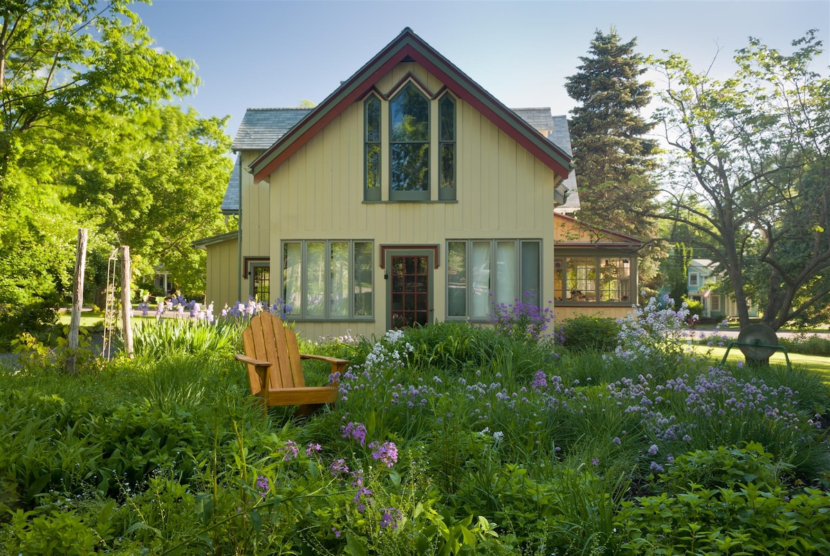 A quaint building is surrounded by lush greenery and vibrant flowerbeds in bloom. A wooden chair is positioned in the foreground, offering a serene view of the garden and the charming structure, which features a mixture of large windows and gabled roofs.