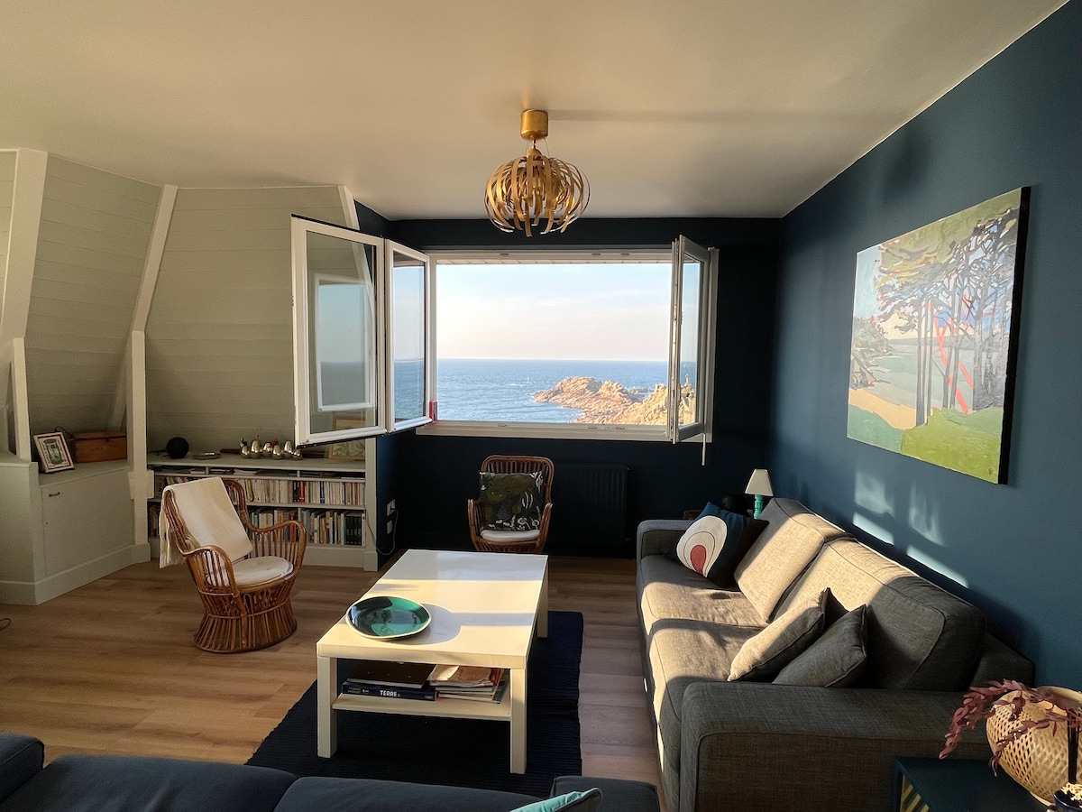 A cozy living room features large windows that frame a sea view, providing ample natural light. The space includes two rattan chairs and a sofa, with a white coffee table at the center. A bookshelf and artwork add character to the room's inviting atmosphere.