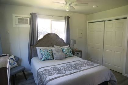 A compact bedroom features a queen bed adorned with decorative pillows and a light grey blanket. A window with sheer curtains allows natural light, while a ceiling fan offers airflow. A small bedside table and a wardrobe with sliding doors complete the space.