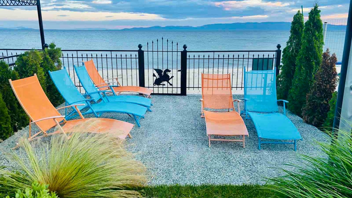 A private outdoor area features six loungers in orange and blue, arranged on a stone patio. Lush greenery lines the space, separating it from the sandy beach just beyond a decorative fence. The sea horizon is visible in the background under a softly lit sky.