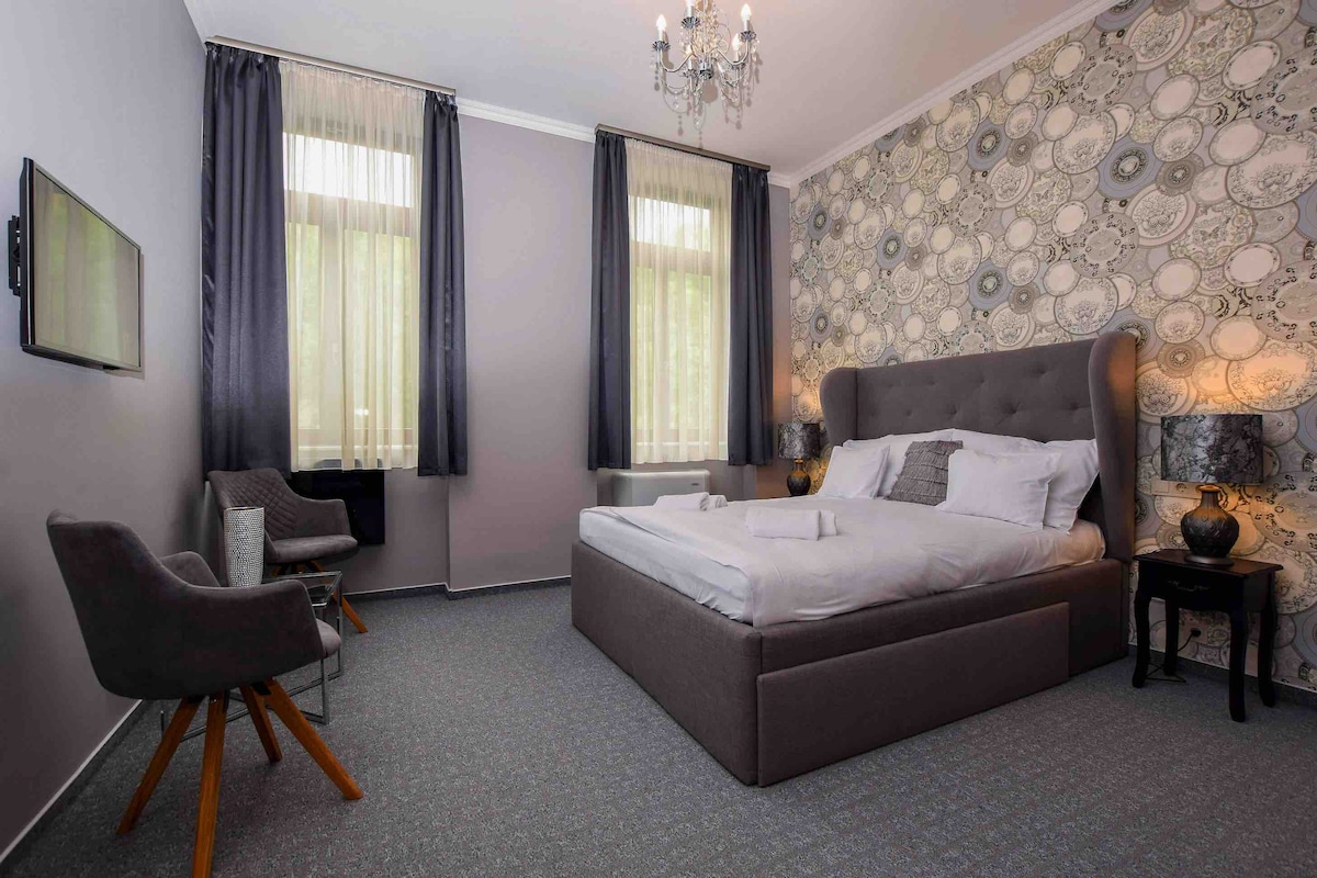 A modern bedroom features a spacious bed with plush bedding, complemented by an elegant tufted headboard. Two armchairs sit beside a decorative table, while large windows dress the room in natural light, framed with dark curtains. A chandelier adds a touch of sophistication.