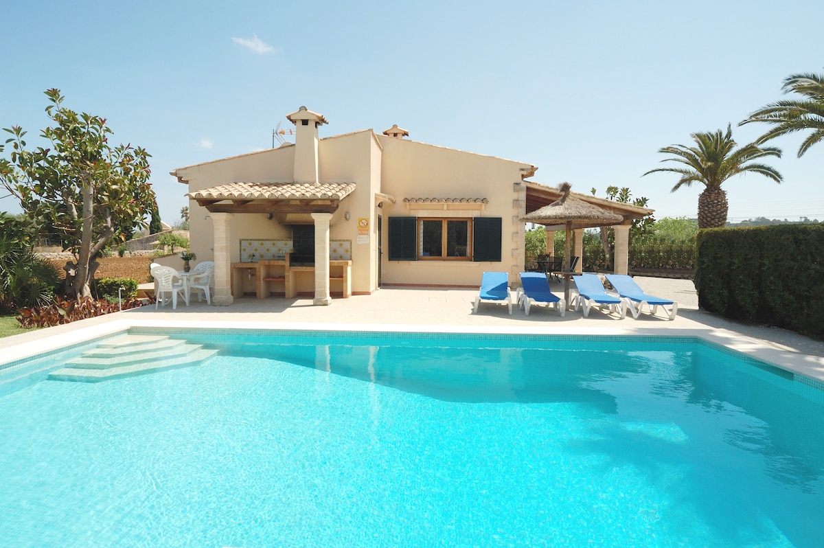 A spacious villa is set beside a clear blue pool, with the house featuring a shaded terrace and outdoor seating. Lush greenery and palm trees surround the area, offering a relaxed outdoor atmosphere. Clear sky and sunlight enhance the inviting scenery.