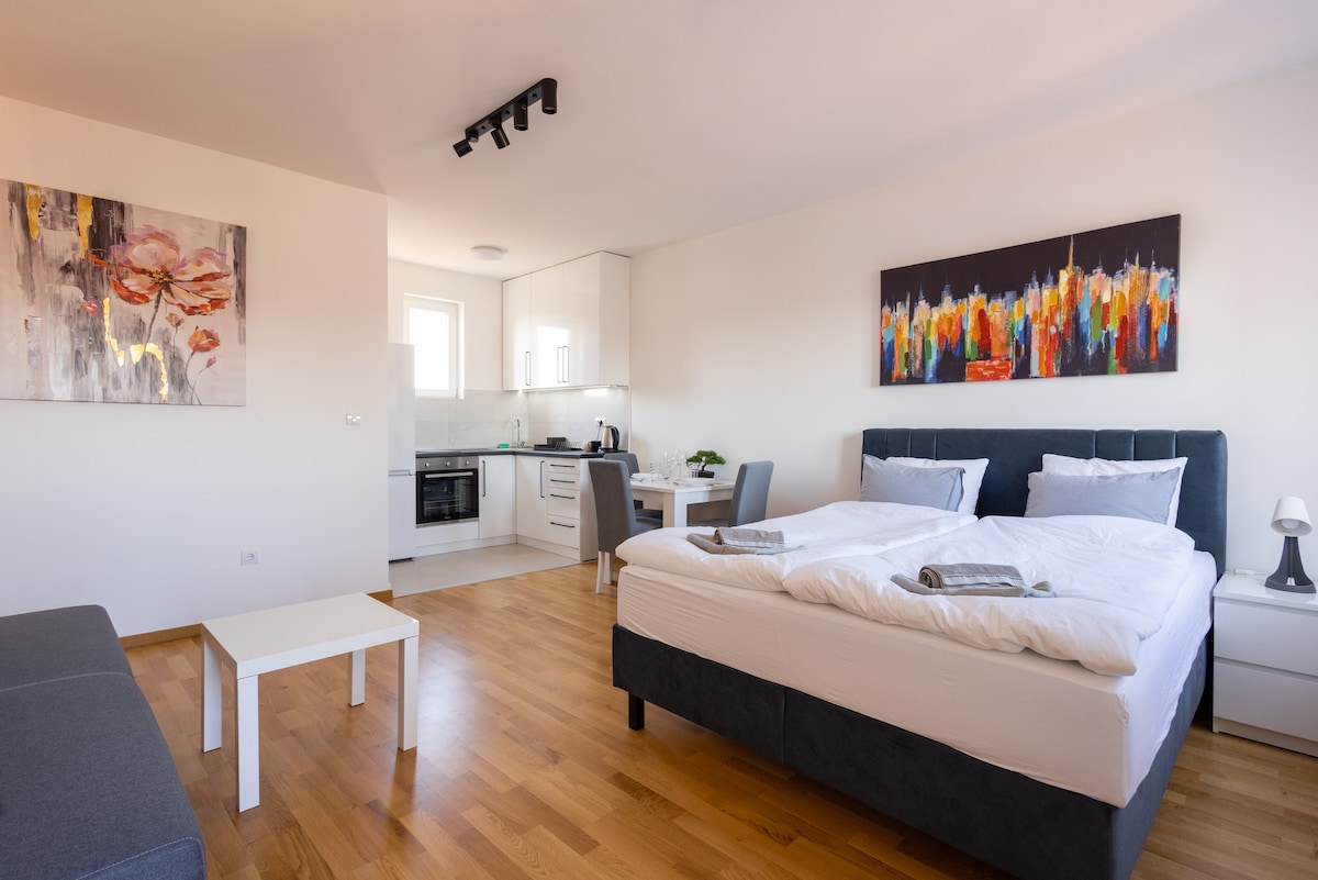 A spacious living area is furnished with a large king bed and a comfortable sofa. A minimalist dining table is positioned near the kitchenette, featuring modern appliances. Art is displayed on the wall, adding color to the room, while light wood flooring enhances the overall brightness.