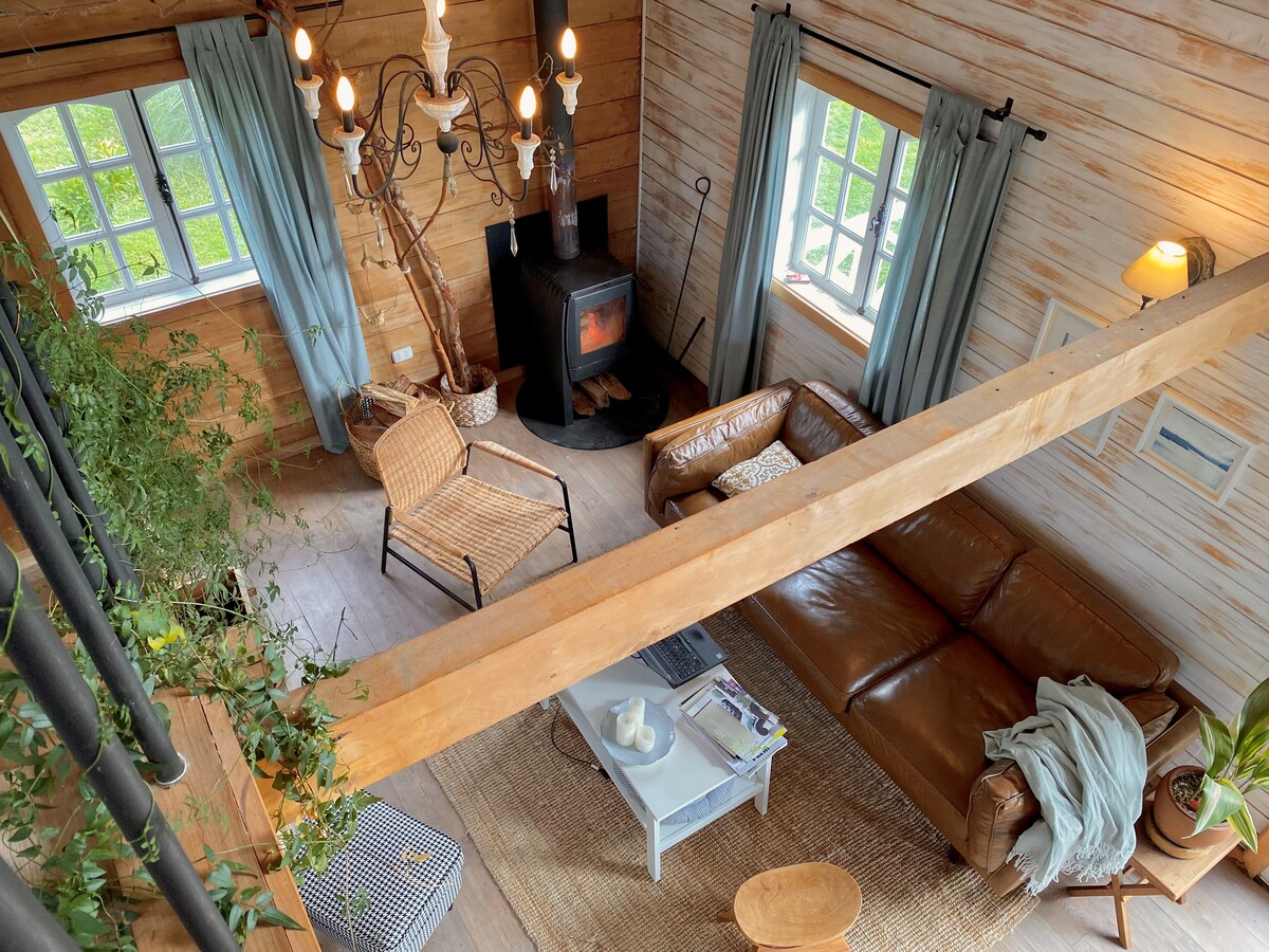 A spacious living area showcases a warm color palette with wooden walls and large windows. Comfortable seating includes a leather sofa and woven chairs. A rustic wood stove and chandelier add character, while soft curtains frame the windows, allowing natural light to brighten the space.