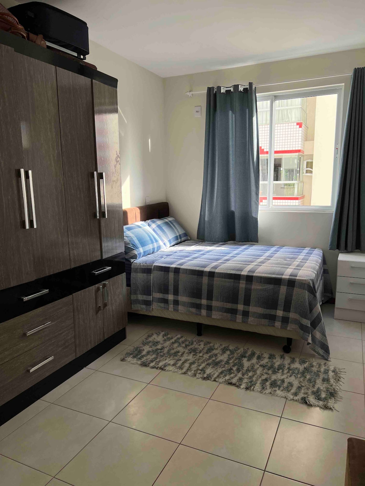 A well-lit bedroom features a spacious bed with a plaid blue and white blanket, positioned against a wall with a window offering natural light. A sturdy wardrobe and a small white nightstand are visible, complementing the room's functionality. Light-coloured tiles enhance the bright atmosphere.