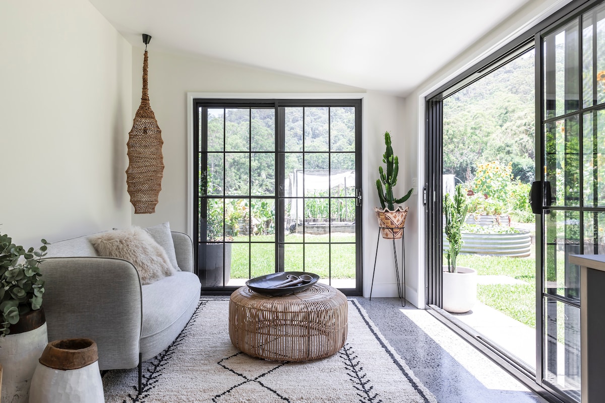 A spacious living area features large black-framed windows providing views of the lush garden. A cozy grey sofa is paired with a round natural fiber coffee table on a patterned rug. Greenery is visible outside, enhancing the connection with nature.