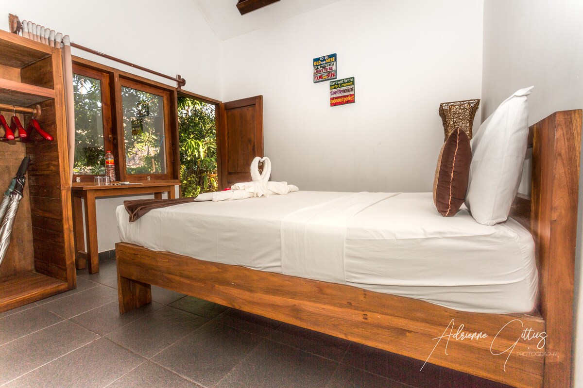 A cozy sleeping area features a queen size bed with crisp white linens arranged neatly. Natural light enters through wooden-framed windows, revealing a garden view. A storage unit stands nearby, including decorative items and a pair of red shoes. Wall art adds a subtle charm.