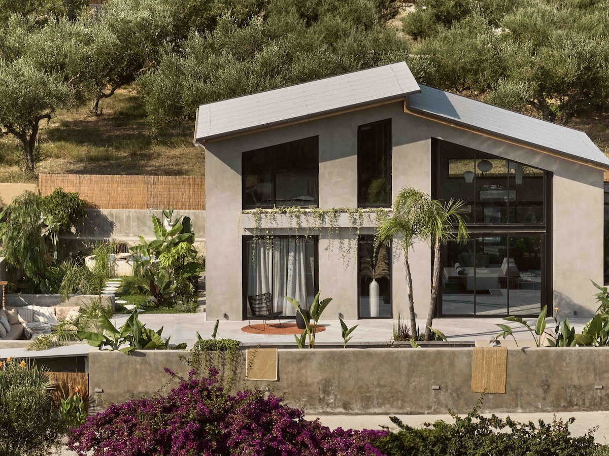 A modern residence with large glass windows is set amidst lush greenery and vibrant plants. The exterior features a blend of earthy tones and sleek lines, complemented by a cozy seating area. Tropical landscaping surrounds the property, creating a serene atmosphere.
