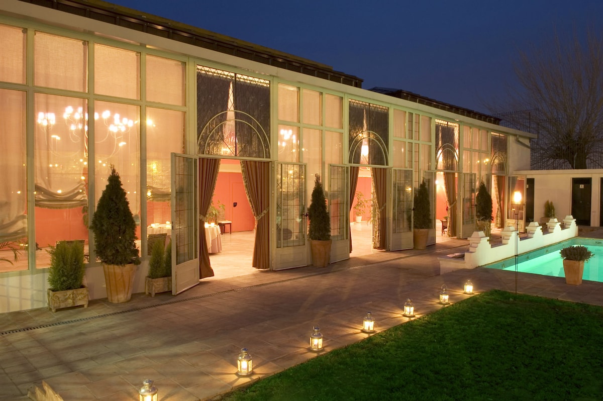 An elegantly designed outdoor area features large glass doors opening to a stylish interior, illuminated by chandeliers. Soft, warm lighting sets a tranquil mood, while lanterns line the path beside a swimming pool, enhancing the serene atmosphere. Lush greenery and potted plants create a welcoming environment.