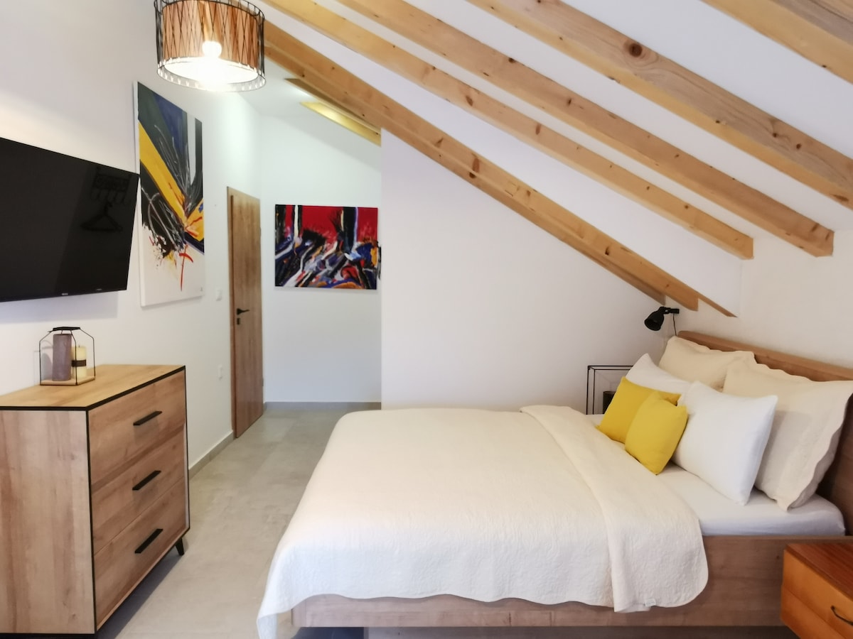 A spacious bedroom features a king size double bed dressed in neutral linens, complemented by yellow accent pillows. Decorated with modern artwork, the room includes a dresser and a flat-screen TV mounted on the wall, all beneath a sloped wooden ceiling.