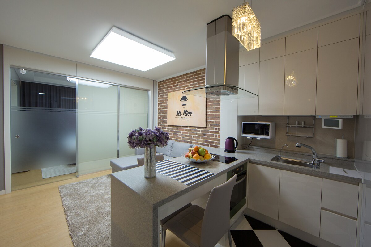 A modern kitchen features sleek cabinetry and a large island with a decorative fruit bowl. Subtle lighting from a stylish chandelier illuminates the space. A cozy seating area is visible in the background, complemented by a contemporary design and warm tones.