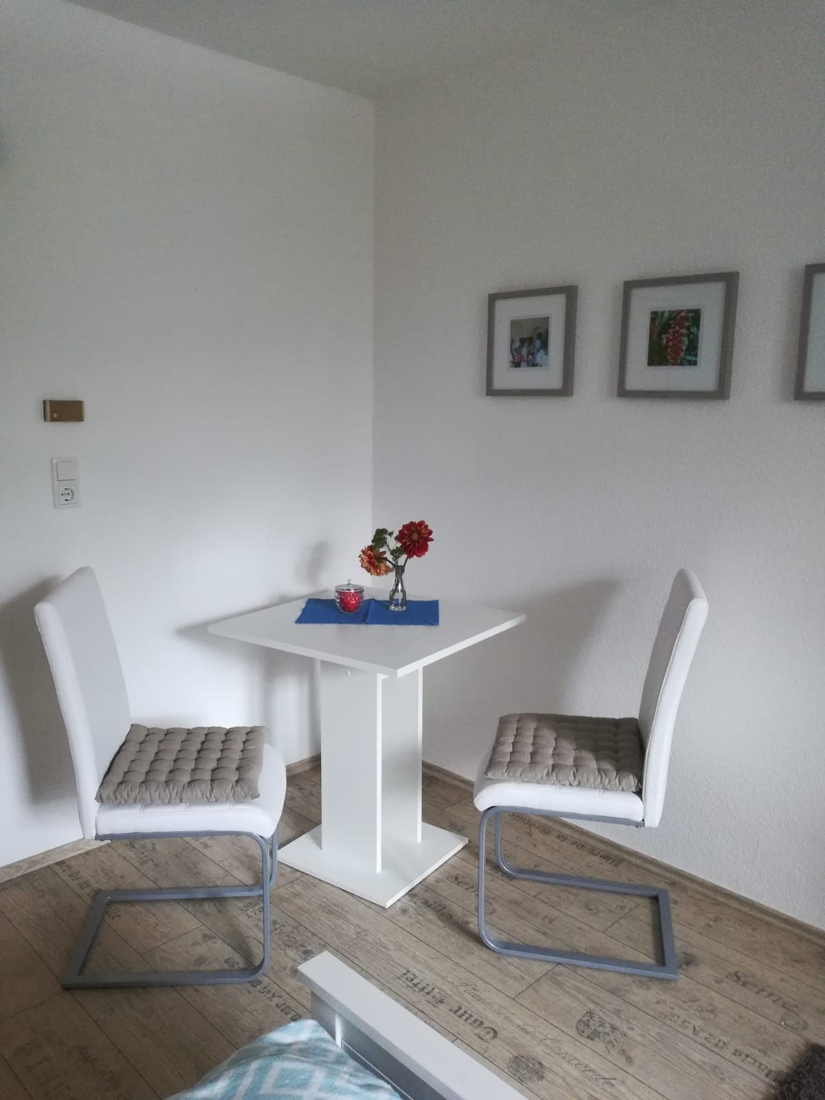 A small dining area is depicted, featuring a square white table complemented by two cushioned chairs. A decorative arrangement of flowers sits atop the table. Wall-mounted frames in a neutral tone display images, contributing to the overall minimalist aesthetic.