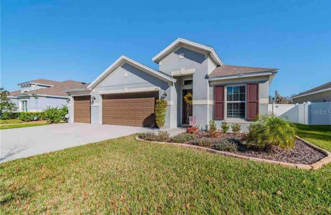 The exterior of a single-story home is presented, featuring a two-car garage and a welcoming front porch. Neatly manicured landscaping includes shrubs and ornamental plants, while the lawn is well-maintained. The home is surrounded by clear blue skies, enhancing its inviting appearance.