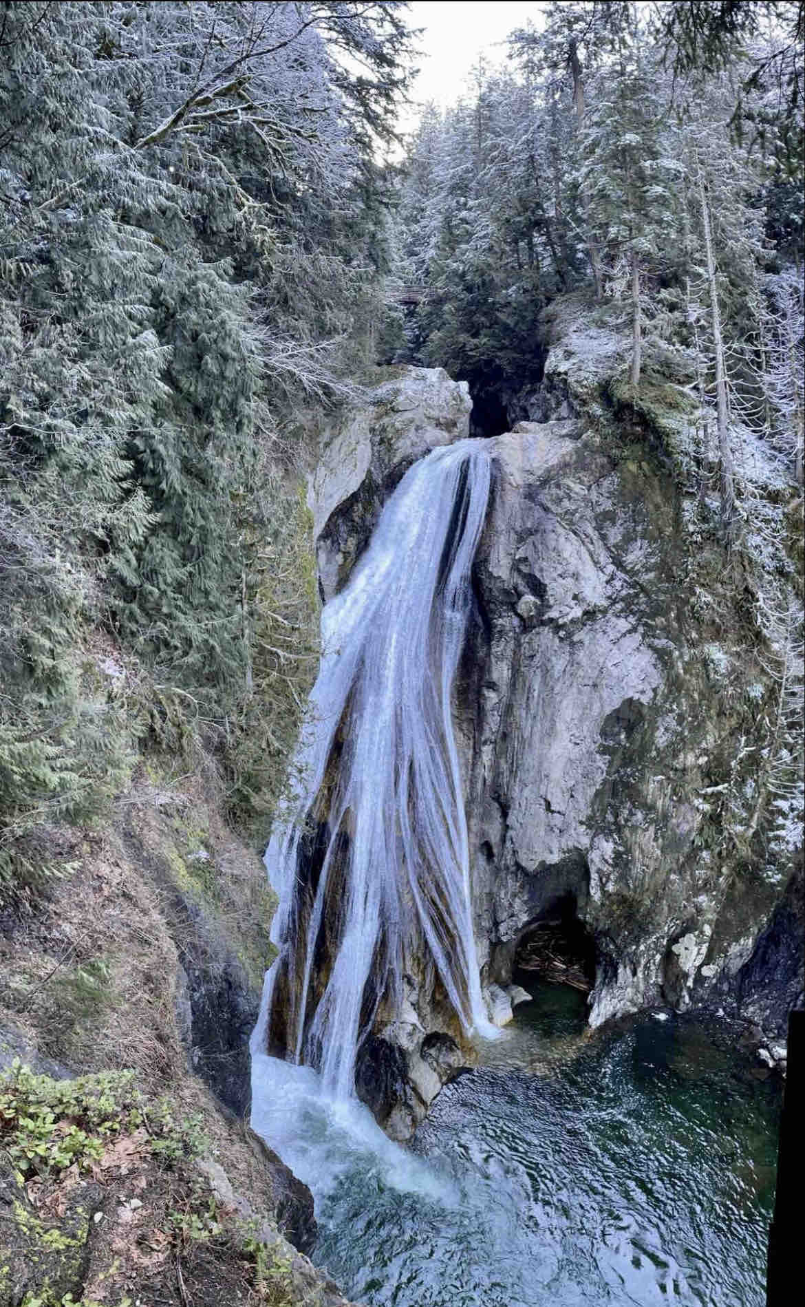 A flowing waterfall cascades down a rocky cliff, surrounded by dense evergreen trees and rocky terrain. The water creates a mist at its base, where it meets a calm pool. Frosty conditions are visible on the surrounding foliage, indicating a winter environment.