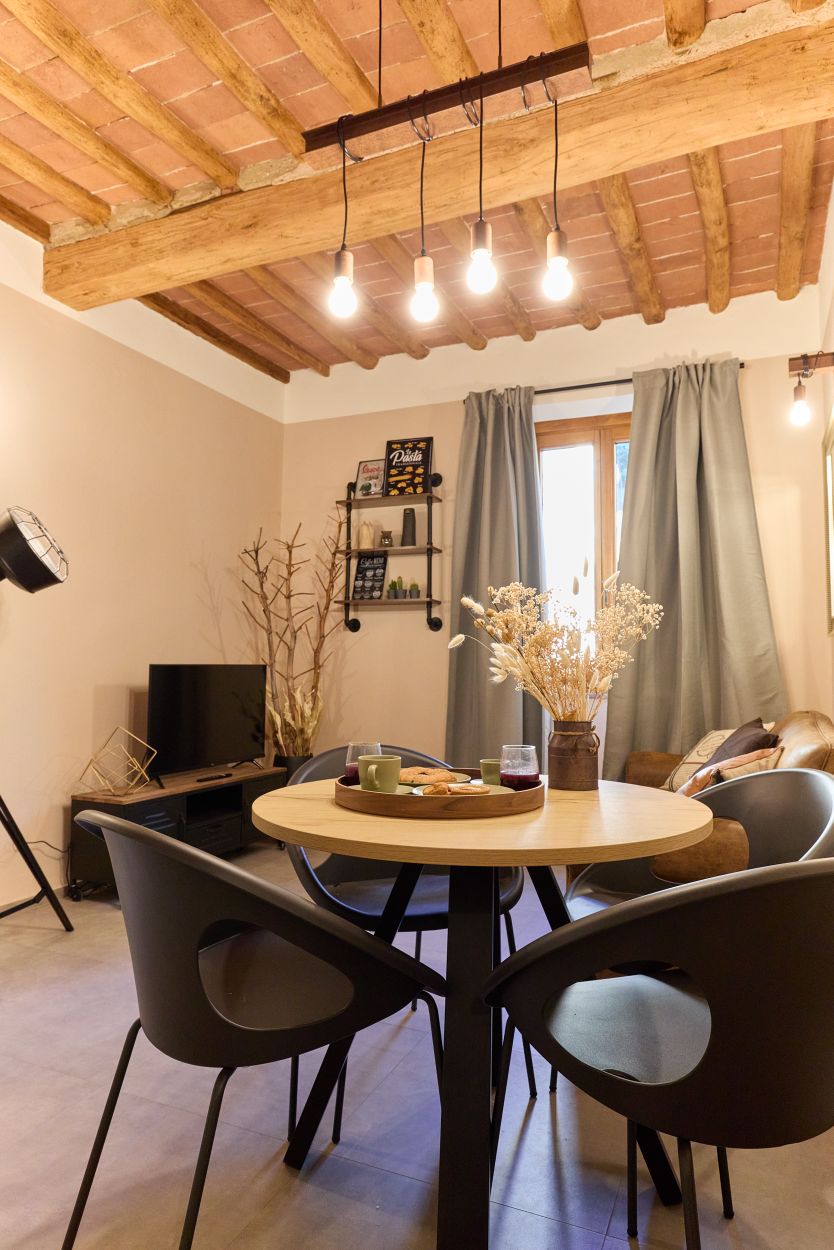 A cozy dining area features a round table surrounded by four modern chairs. Above, warm lighting hangs from a rustic ceiling with exposed beams. A small shelf displays decorative items, while a television is positioned across from a comfortable seating area, adding to the inviting atmosphere.