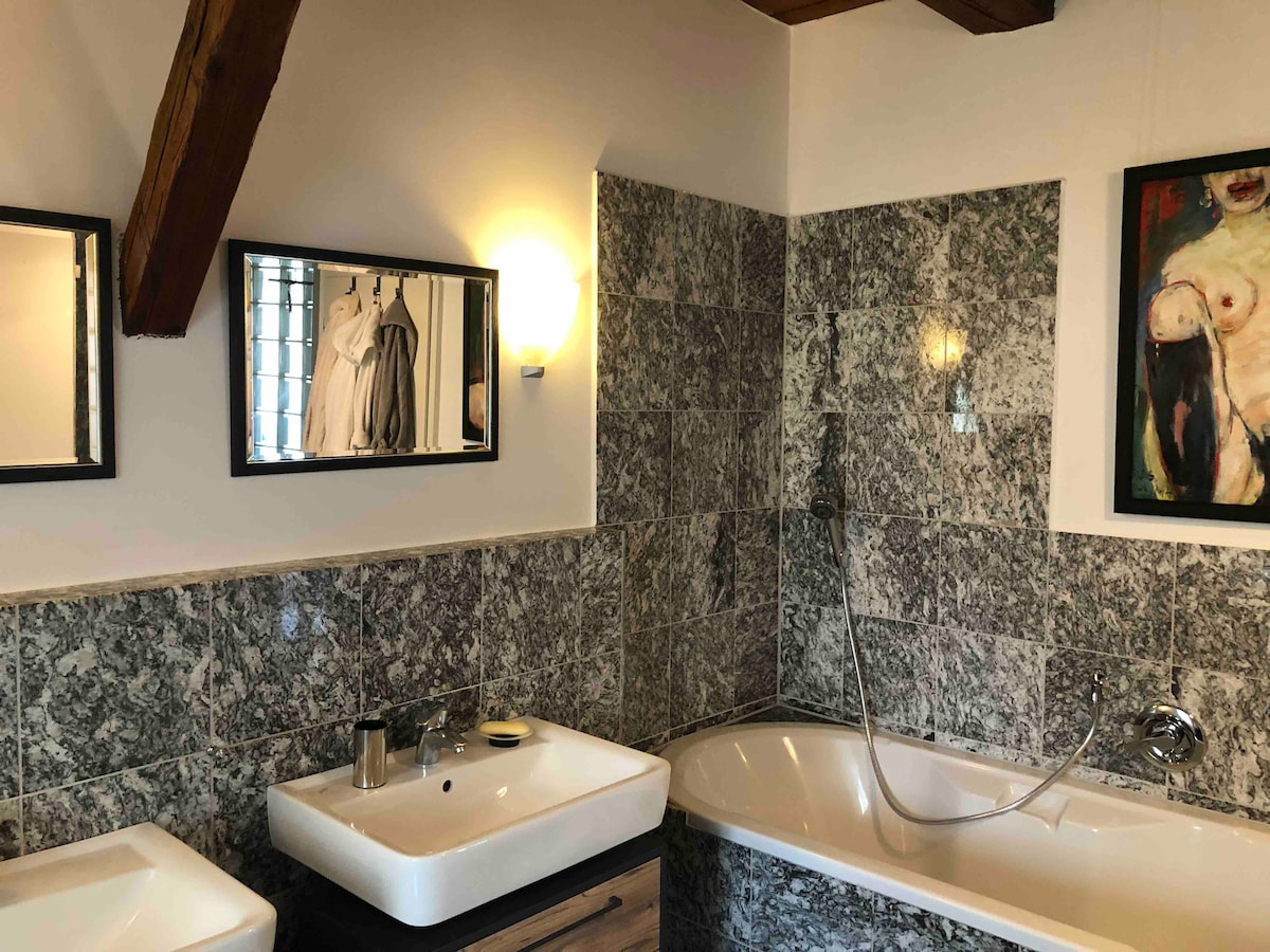 The bathroom features a marble-tiled wall, highlighted by modern lighting. A spacious bathtub is positioned alongside two sinks with sleek mirrors above them. Artwork decorates one wall, adding a touch of elegance to the space.