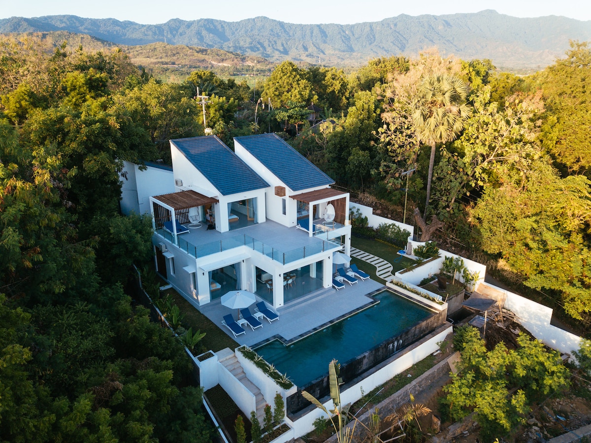 An aerial view of a modern villa surrounded by lush greenery and mountains in the background. The villa features a large pool in the foreground and multiple outdoor seating areas. The clean architectural lines highlight the integration of indoor and outdoor spaces.