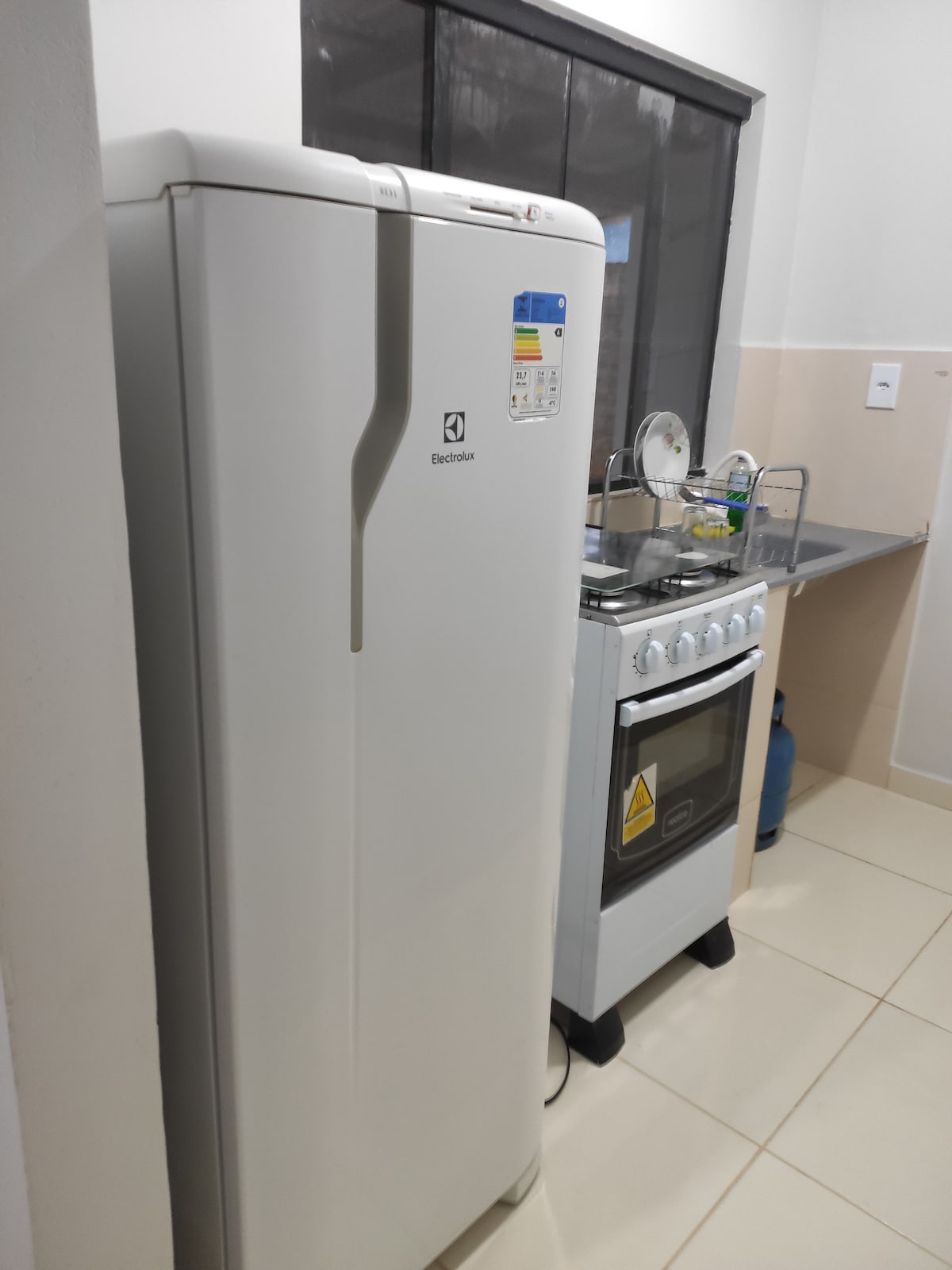 A functional kitchen features a white refrigerator and a compact stove with an oven. Counter space is provided for meal preparation, and a small window allows for natural light. Various utensils can be seen on the countertop, contributing to a practical cooking environment.